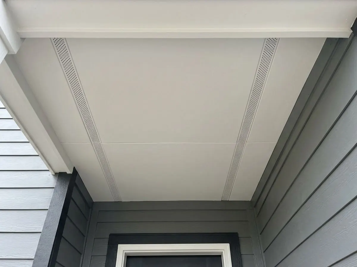 Looking up at a deep roof overhang finished with smooth white flat soffit panels and small vented sections, above grey lap siding.