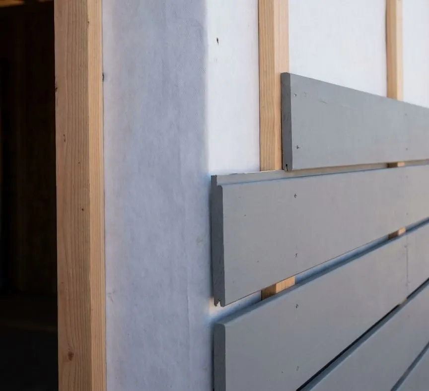 Grey horizontal boards fastened to vertical wood furring strips over white house wrap, leaving an open drainage gap behind the cladding.